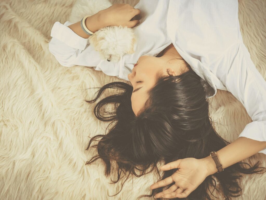 白いシャツを着た女性が毛布の上で小さな犬を抱きながら安らかに眠っている様子。 深いリラックスと安心感を表すシーン。