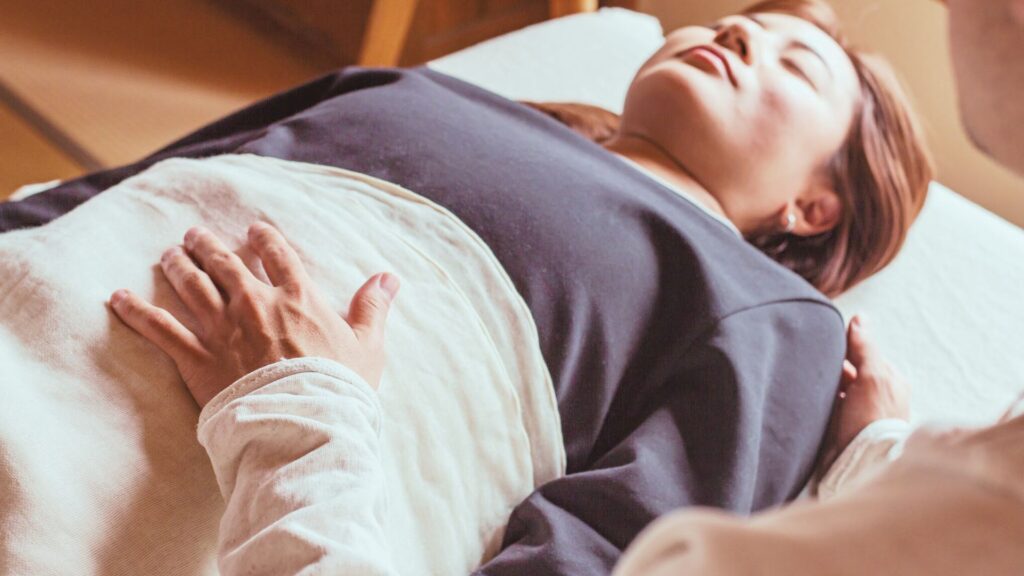 腹部にそっと手を当てる腸もみ(チネイザン)の施術風景。女性がリラックスした姿勢で横になり、やさしいタッチによるチネイザンで腸と自律神経を整えている様子。お腹の不調は食事以前に睡眠にあると思っています。