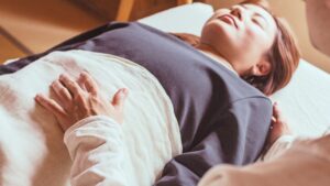 腹部にそっと手を当てる腸もみ（チネイザン）の施術風景。女性がリラックスした姿勢で横になり、やさしいタッチによるチネイザンで腸と自律神経を整えている様子。お腹の不調は食事以前に睡眠にあると思っています。