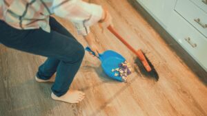 床に散らばったゴミをほうきとちりとりで掃除する様子、家事を行う女性の手元、家庭内の掃除風景、家事ストレスや日常生活のイメージ